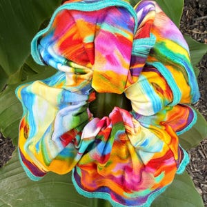 May include: A fabric hair scrunchie featuring a vibrant, multi-colored abstract pattern with swirls of bright yellow, orange, red, blue, and purple. The circular accessory has a gathered, ruffled texture and is finished with a teal green stitched edge.