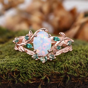 May include: A rose gold ring featuring a central oval-shaped iridescent opal stone. The band is designed with intricate metal leaf motifs and is accented with small, marquise-cut green stones embedded along the branches surrounding the opal.