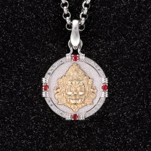 Może przedstawiać: Srebrny łańcuszek z okrągłym wisiorkiem przedstawiającym złotą boginię o groźnym wyrazie twarzy. Wisiorek jest otoczony srebrną obręczą z małymi czerwonymi kamieniami szlachetnymi.