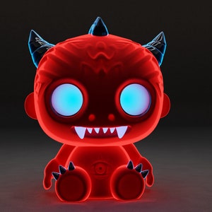 May include: A red, cartoon-style devil with black horns and glowing blue eyes. The devil is sitting with its legs crossed and has a toothy grin.