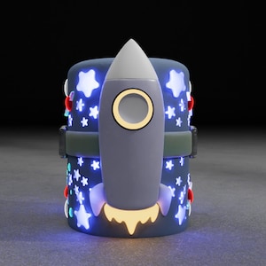 May include: A blue and white rocket-shaped nightlight with a yellow flame and stars on the base. The nightlight is lit up with blue and yellow lights.