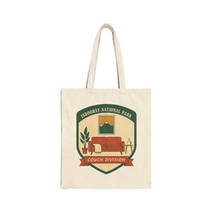 May include: Cream-colored tote bag with a graphic of a living room scene inside a green and tan badge. The badge reads "INDOORSY NATIONAL PARK" and "COUCH DIVISION". The graphic includes a red couch, plant, and coffee table.