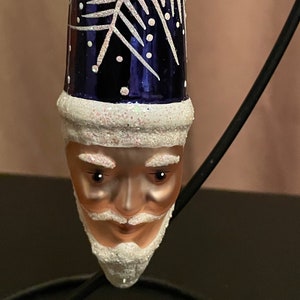 May include: A blue glass Christmas ornament shaped like a Santa Claus head with a white snowflake design and a white beard and mustache.