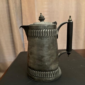 May include: Antique silver-colored coffee pot with a black handle. The pot features decorative vertical ridges and a detailed lid with a small finial. The spout is curved, and the handle is attached with decorative metal accents.