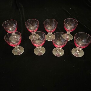May include: Eight pink-tinted glass goblets with clear stems and bases are arranged on a dark surface. The bowls of the glasses have vertical etched lines. The goblets are likely for serving wine or other beverages.