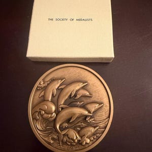 May include: A bronze-colored medal featuring a relief of dolphins leaping from waves. The medal is round and has the text "THE SOCIETY OF MEDALISTS" on the accompanying cream-colored box.