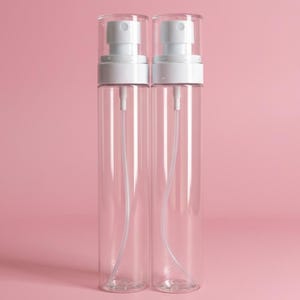 May include: Two clear, cylindrical spray bottles with white spray nozzles and caps. The bottles have a long, thin, clear tube inside. The background is a solid pink color.