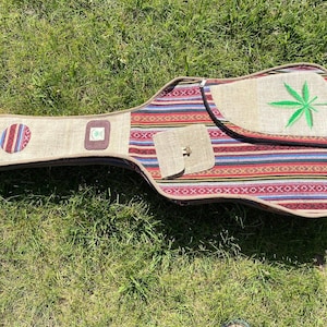 May include: A guitar case with a hemp-like fabric exterior. The case features a colorful striped pattern and a green embroidered cannabis leaf. It has a small pocket with a metal clasp and a circular patch.