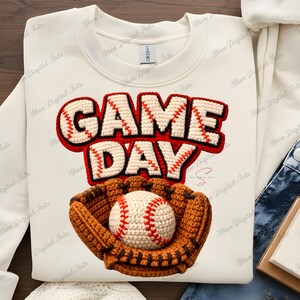 May include: Cream-colored sweatshirt with a baseball-themed design. The words "GAME DAY" are in a 3D stitched style, with a baseball and mitt below. The baseball is white with red stitching, and the mitt is brown.