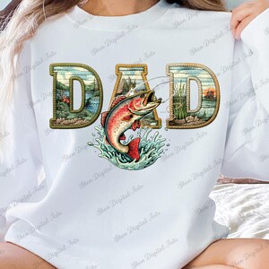 Puede incluir: Sudadera blanca con la palabra "DAD" en una fuente decorativa con un tema de pesca. Las letras están rellenas de escenas de paisajes, y un pez saltando está en el centro. El diseño incluye salpicaduras de agua y una línea de pesca.