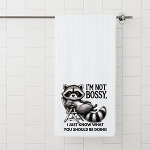 May include: White bath towel with a raccoon illustration. The raccoon is sitting in a chair and the text reads "I'M NOT BOSSY, I JUST KNOW WHAT YOU SHOULD BE DOING". The towel is hanging on a metal bar.