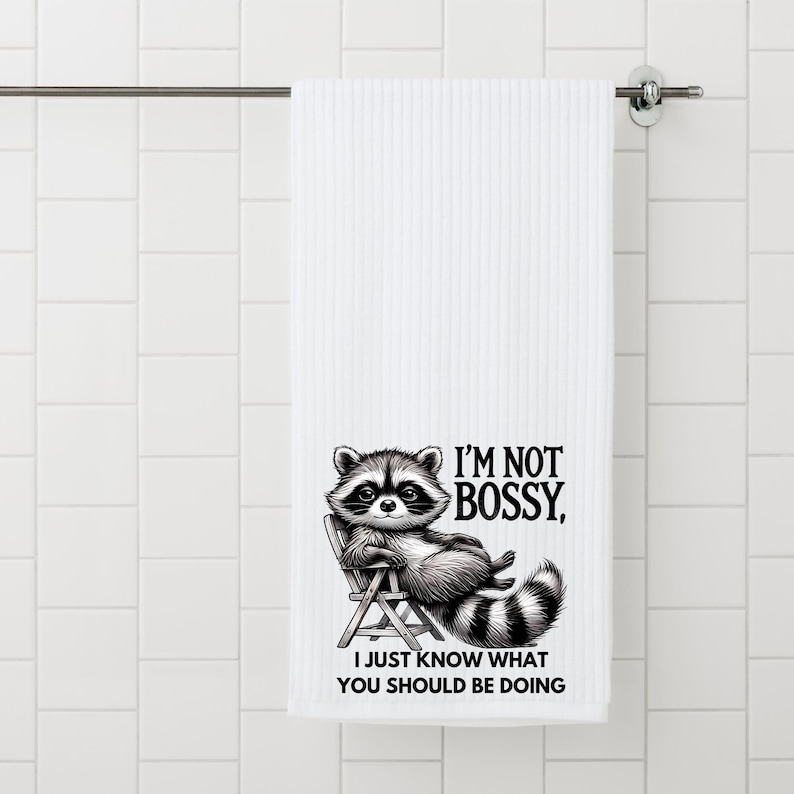 May include: White bath towel with a raccoon illustration. The raccoon is sitting in a chair and the text reads "I'M NOT BOSSY, I JUST KNOW WHAT YOU SHOULD BE DOING". The towel is hanging on a metal bar.