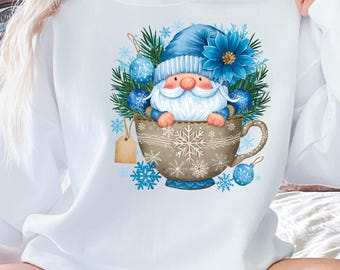 May include: White sweatshirt featuring a whimsical winter gnome design. The gnome, with a blue hat and white beard, is nestled in a decorative teacup adorned with snowflakes and blue ornaments. A tag hangs from the cup, and snowflakes are scattered around.