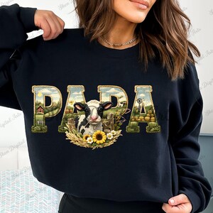 Peut inclure: Sweat-shirt noir avec le mot "PAPA" dans une police décorative. Les lettres sont remplies d'une scène de ferme, et une vache est au centre, entourée de tournesols. Le sweat-shirt est de style décontracté.