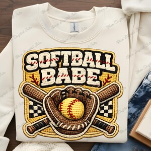 Può includere: Felpa color crema con un design a tema softball. Il design include le parole "SOFTBALL BABE" in uno stile cucito, una palla da softball gialla, un guanto marrone e mazze incrociate. Il design è delineato in nero e giallo.
