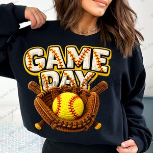 May include: Black sweatshirt with a baseball-themed design. The words "GAME DAY" are in white with yellow and red accents, above a baseball glove holding a yellow softball. Two brown baseball bats are behind the glove.