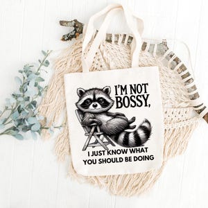 May include: Cream-colored tote bag featuring a raccoon illustration lounging in a chair. The raccoon is black and white, with the text "I'M NOT BOSSY, I JUST KNOW WHAT YOU SHOULD BE DOING" printed in black. The bag has two cream-colored handles.
