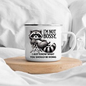 May include: White enamel mug with a black and white raccoon illustration. The raccoon is lounging in a chair, with the text "I'M NOT BOSSY, I JUST KNOW WHAT YOU SHOULD BE DOING". The mug has a silver rim and a white handle.