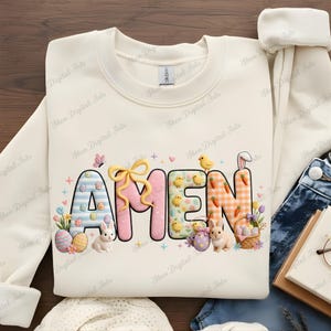 Può includere: Felpa color crema con la scritta "AMEN" in lettere colorate a tema pasquale. Le lettere sono decorate con uova di Pasqua, coniglietti, pulcini, carote e un fiocco. La felpa è a girocollo.