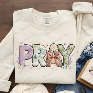 Può includere: Felpa color crema con la parola "PRAY" in un carattere decorativo e colorato. La lettera "P" presenta croci e lavanda, la "R" ha un fiocco e la "Y" ha accenti floreali. Sono presenti anche una colomba e un'aureola.