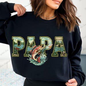Puede incluir: Sudadera negra con la palabra "PAPA" en un diseño de paisaje verde y azul. Un pez rojo salta fuera del agua en el centro de la palabra "PAPA".