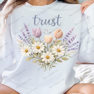 Può includere: Felpa azzurra con la scritta "trust" in un font glitterato sopra un disegno floreale. La composizione floreale comprende margherite, tulipani e lavanda in tenui colori pastello.