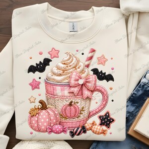 May include: A cream-coloured sweatshirt with a Halloween-themed design. A pink and white gingham mug is filled with whipped cream and a candy cane. The design includes pumpkins, bats, stars, and candies in pink, black, and orange.