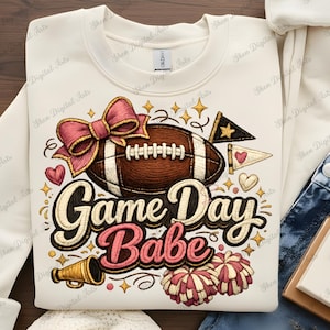 May include: Cream-colored sweatshirt with a football and the text "Game Day Babe." The design includes a pink bow, hearts, stars, a megaphone, and pom-poms. The football is brown and white.