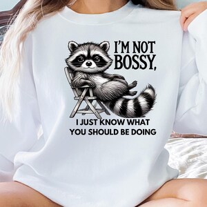 May include: White sweatshirt featuring a detailed illustration of a raccoon lounging in a chair. The raccoon has black and white fur and is smiling. Text reads "I'M NOT BOSSY, I JUST KNOW WHAT YOU SHOULD BE DOING."