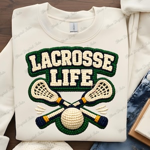Puede incluir: Sudadera crema con un diseño verde y azul marino. El diseño presenta las palabras "LACROSSE LIFE" sobre un gráfico de palos y pelotas de lacrosse cruzados. El gráfico está en un estilo texturizado y cosido.