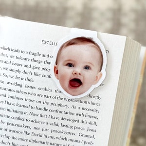 May include: A white bookmark featuring a smiling baby's face is placed on the pages of a book. The bookmark is oval-shaped and the baby has an open mouth. The book is open to a page with text.