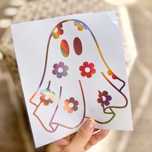 K&ouml;nnte beinhalten: Ein wei&szlig;er Vinyl-Aufkleber mit einem Geist mit regenbogenfarbenem, irisierendem Finish. Der Geist hat ein einfaches, cartoonartiges Design und ist mit kleinen Blumenformen verziert.