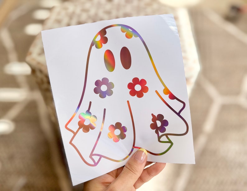 K&ouml;nnte beinhalten: Ein wei&szlig;er Vinyl-Aufkleber mit einem Geist mit regenbogenfarbenem, irisierendem Finish. Der Geist hat ein einfaches, cartoonartiges Design und ist mit kleinen Blumenformen verziert.