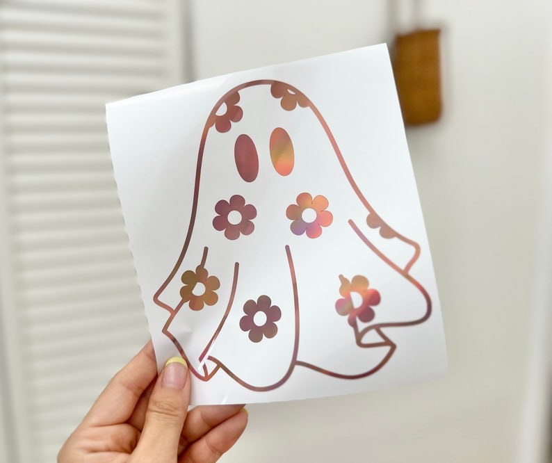 K&ouml;nnte beinhalten: Ein wei&szlig;er Vinyl-Aufkleber mit einem rosa schimmernden Geist mit Blumen. Der Geist ist schwarz umrandet und hat ein einfaches Gesicht mit zwei Augen.