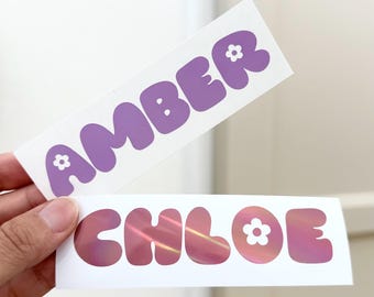 May include: Two rectangular stickers with names. The top sticker reads "AMBER" in purple bubble letters with small flower accents. The bottom sticker says "CHLOE" in pink holographic bubble letters, also with flower accents. Both stickers are on a white background.