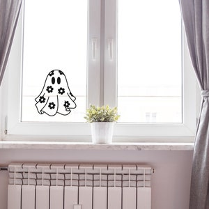K&ouml;nnte beinhalten: Ein wei&szlig;er Geist mit schwarzen Augen und einem Blumenmuster auf seinem Laken befindet sich an einem Fenster. Der Geist befindet sich vor einem wei&szlig;en Fensterrahmen und grauen Vorh&auml;ngen.