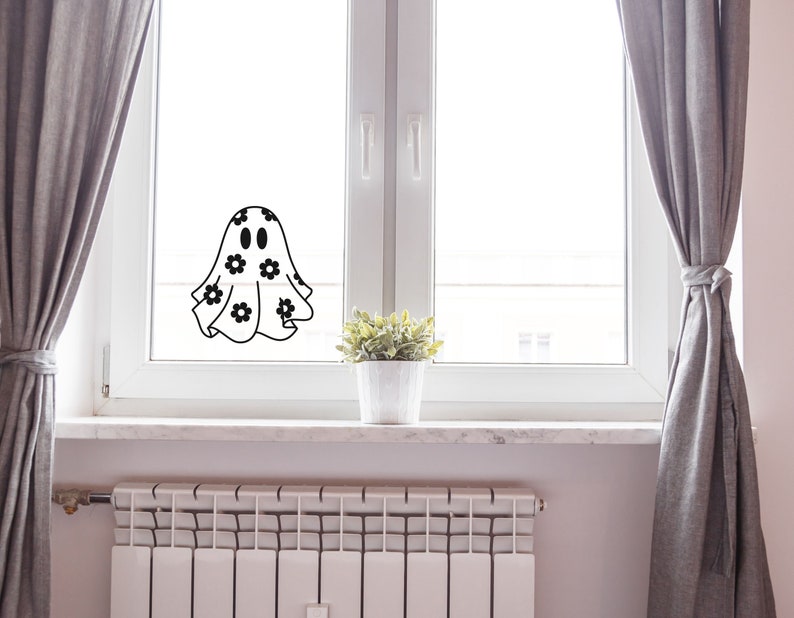 K&ouml;nnte beinhalten: Ein wei&szlig;er Geist mit schwarzen Augen und einem Blumenmuster auf seinem Laken befindet sich an einem Fenster. Der Geist befindet sich vor einem wei&szlig;en Fensterrahmen und grauen Vorh&auml;ngen.