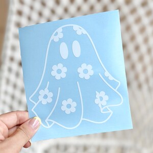 K&ouml;nnte beinhalten: Ein wei&szlig;er Vinyl-Aufkleber mit einem Geist mit Blumenmuster. Der Geist hat zwei Augen und ein einfaches L&auml;cheln. Der Aufkleber befindet sich auf einem hellblauen Hintergrund.