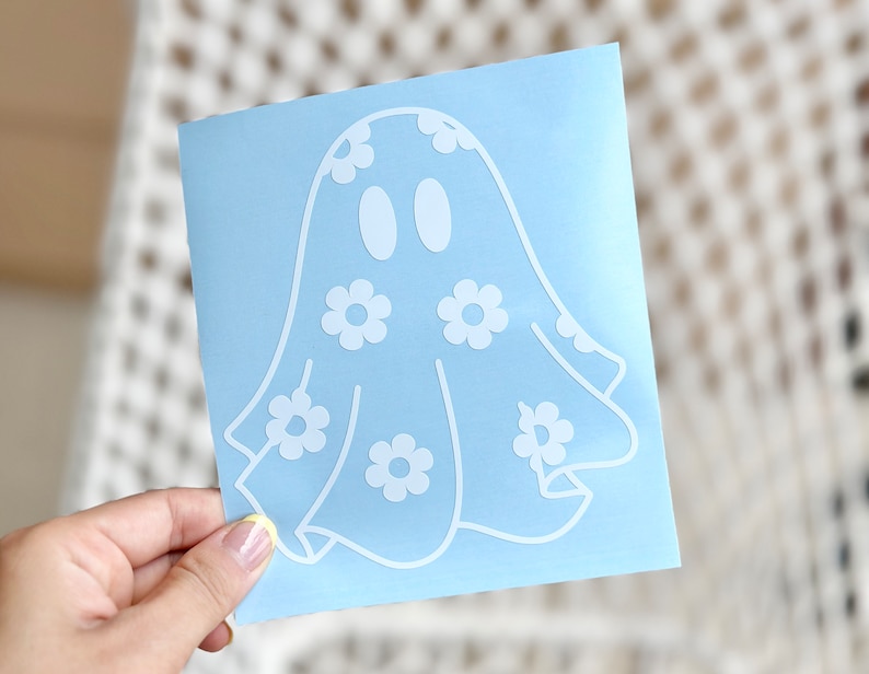 K&ouml;nnte beinhalten: Ein wei&szlig;er Vinyl-Aufkleber mit einem Geist mit Blumenmuster. Der Geist hat zwei Augen und ein einfaches L&auml;cheln. Der Aufkleber befindet sich auf einem hellblauen Hintergrund.