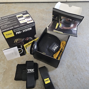 May include: TRX PRO4 SYSTEM suspension training kit. The black box contains a black mesh bag, yellow straps, and black suspension straps. The box has the TRX logo and the words "MAKE YOUR BODY YOUR MACHINE".