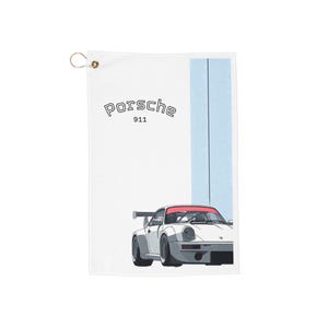 May include: White towel featuring a Porsche 911 illustration. The towel has a light blue vertical stripe on the right side and a gold grommet at the top. The Porsche logo and "911" are printed in black.