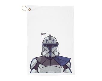 May include: White golf towel featuring a stylized illustration of a clone trooper helmet and armor in shades of gray and blue. The towel has a brass grommet for hanging. Ideal for Star Wars fans.