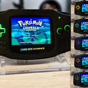 Puede incluir: Una consola Game Boy Advance SP negra con retroiluminaci&oacute;n verde. La consola muestra el juego Pokemon Emerald Version. La consola est&aacute; en un soporte de acr&iacute;lico transparente.