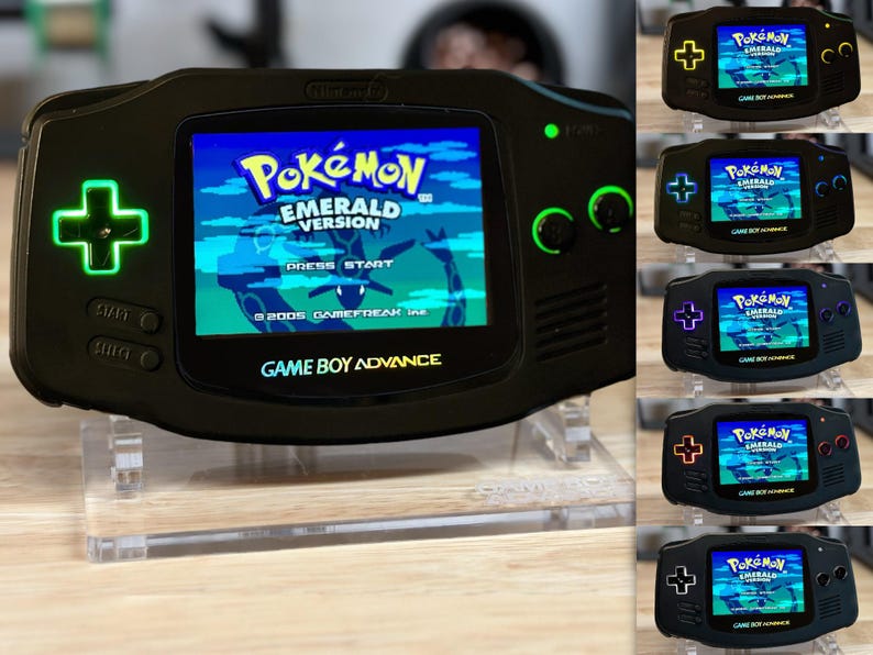 Puede incluir: Una consola Game Boy Advance SP negra con retroiluminaci&oacute;n verde. La consola muestra el juego Pokemon Emerald Version. La consola est&aacute; en un soporte de acr&iacute;lico transparente.