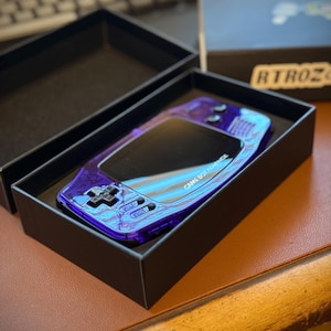 Puede incluir: Una consola Game Boy Color morada en una caja negra. La consola tiene una carcasa transparente morada con un acabado azul iridiscente. La caja tiene el texto "GAME BOY COLOR" impreso en ella.
