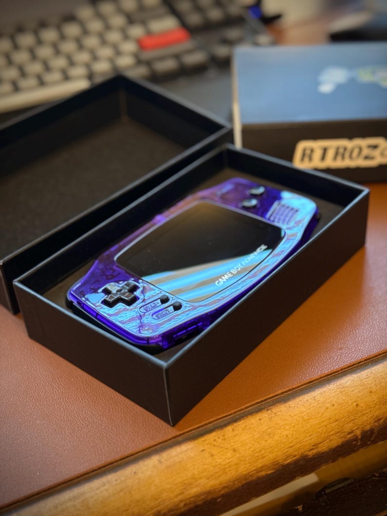 Puede incluir: Una consola Game Boy Color morada en una caja negra. La consola tiene una carcasa transparente morada con un acabado azul iridiscente. La caja tiene el texto "GAME BOY COLOR" impreso en ella.