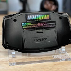 Puede incluir: Una consola de videojuegos port&aacute;til Game Boy Advance negra con una pegatina de c&oacute;digo de barras de colores arco&iacute;ris en la parte posterior. La consola est&aacute; en un soporte de acr&iacute;lico transparente con las palabras "Game Boy Advance" impresas en la base.