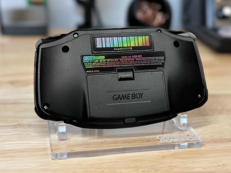 Puede incluir: Una consola de videojuegos port&aacute;til Game Boy Advance negra con una pegatina de c&oacute;digo de barras de colores arco&iacute;ris en la parte posterior. La consola est&aacute; en un soporte de acr&iacute;lico transparente con las palabras "Game Boy Advance" impresas en la base.
