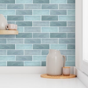 May include: A kitchen backsplash with a blue and gray tile pattern. The tiles are arranged in a brick pattern. There is a white pitcher and a small white jar on a wooden coaster in the foreground.