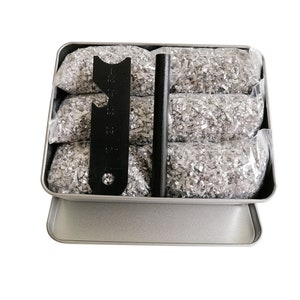 May include: A silver metal tin with a black fire starter, a black bottle opener, and five small bags of silver tinder. The tin is closed and the bags are stacked inside.